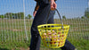 Person holding a yellow basket filled with apples in an outdoor setting.